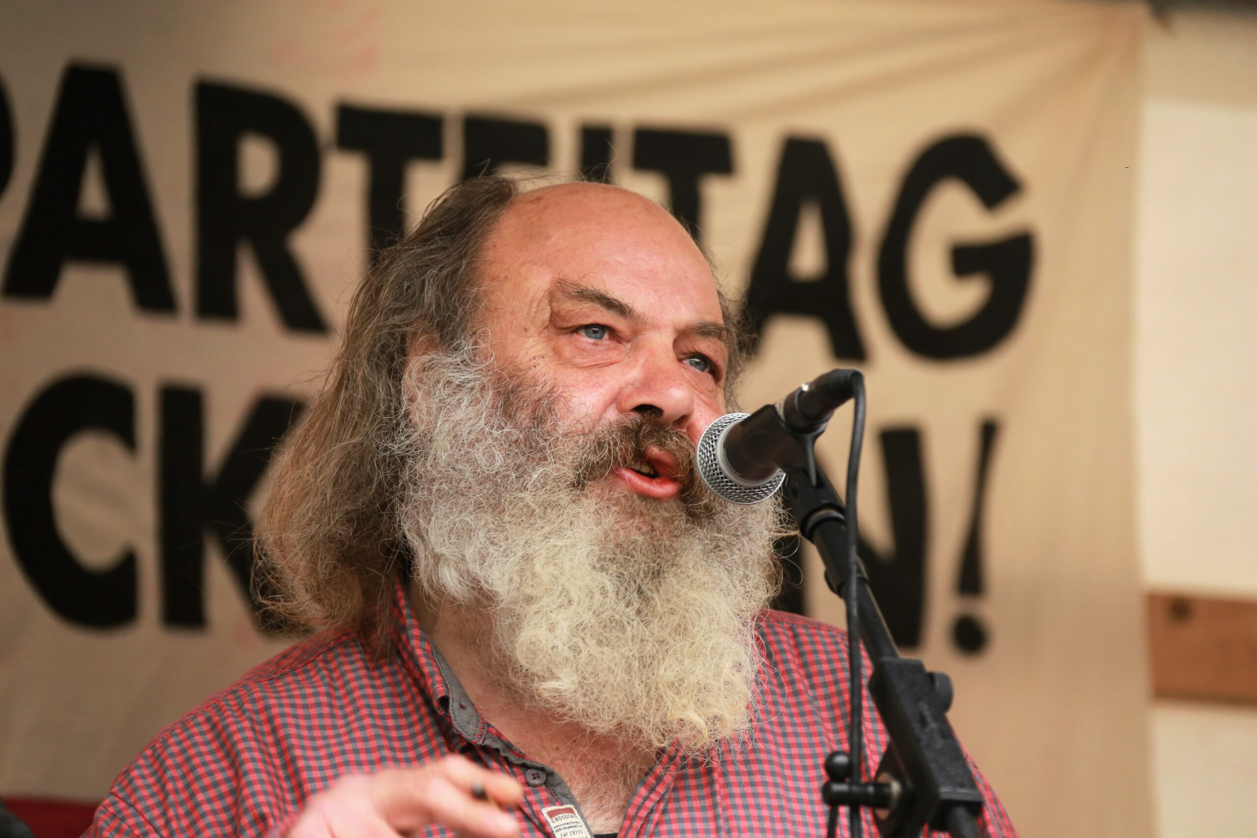 Foto des Pfarrers Lothar König. Zu sehen ein Mann mit langen Haaren und grauem Vollbart im karierten Hemd vor einem Mikrofon.